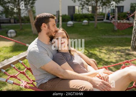 A man and woman enjoy a tranquil moment together on a red hammock in a sunny park. Both looking away the image captures a serene, affectionate moment Stock Photo