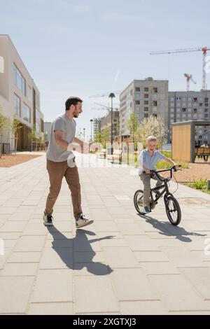 A joyful scene in an urban park where a father, looking at his son, is teaching him how to ride a bicycle. The son, looking away towards the path and Stock Photo