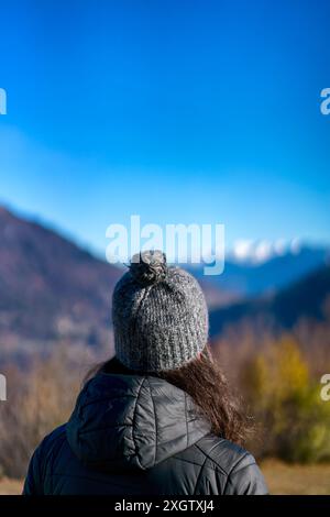 A woman, seen from behind, gazes at a distant range of snow-capped mountains under a clear blue sky, her hair peeking out from under a knitted gray be Stock Photo