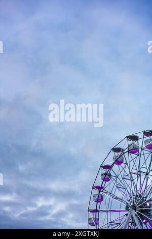 A captivating image of a Ferris wheel set against a moody, overcast sky The Ferris wheel features vivid purple seats, adding a pop of color to the ser Stock Photo