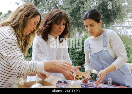 Three women of various ages are gathered at a casual outdoor picnic, engaging with food items served from a takeaway box. They are focused on arrangin Stock Photo