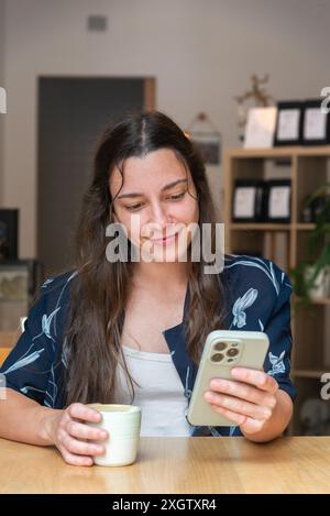 A woman is seen looking down at her smartphone, holding a coffee cup during a break. She appears relaxed and engaged in her mobile device. Stock Photo