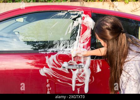 Young woman diligently cleans her red car using a sponge and soapy water, focusing on the details on a sunny day Stock Photo