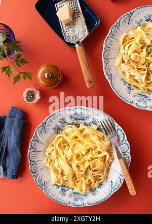 From above, Alfredo pasta dinner with creamy white sauce and herbs served on a red table with pepper mill , parmesan grater and a blue napkin Stock Photo