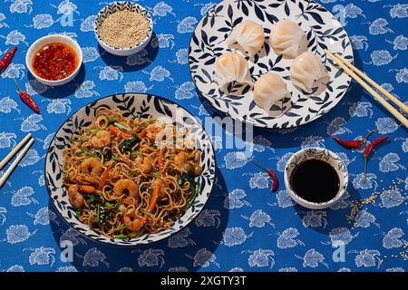 From above, a vibrant display of Chinese food on a blue floral tabletop with shrimp noodles, steamed dumplings in a patterned bowl, and condiments lik Stock Photo