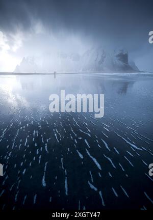 A solitary figure walks on the reflective surface of an Icelandic beach, with majestic snow-covered mountains shrouded in mist in the background. Stock Photo
