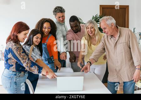 A multiethnic group of male and female professionals excitedly engage over a laptop in a bright coworking space, all looking at the laptop screen and Stock Photo