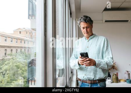Middle-aged Caucasian man in a pastel shirt standing by the window in a modern office space, absorbed in using a smartphone. The setting offers a view Stock Photo