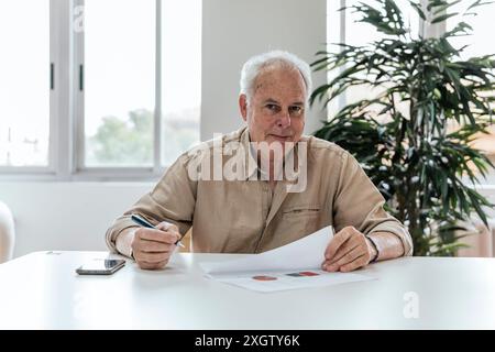 An elderly Caucasian male is seated at a white table in a well-lit office, scrutinizing a document and looking directly at the camera. A smartphone an Stock Photo
