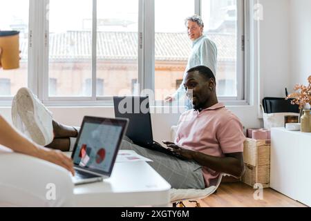 In a sunlit coworking environment, an African American man typing on a laptop looks at his screen while a senior Caucasian man stands in the backgroun Stock Photo
