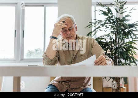 A thoughtful elderly Caucasian man reviews documents intently at a white table, showing signs of stress, in a well-lit room with lush indoor plants. H Stock Photo