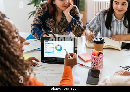 A multiracial cropped group of professionals collaborates over a budget analysis in a well-lit coworking space. The focus is on a digital tablet displ Stock Photo