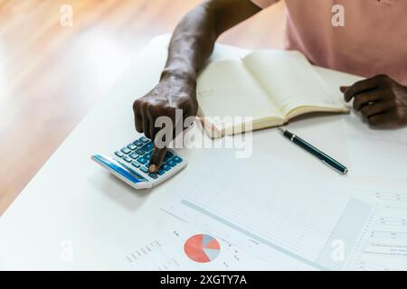 Cropped unrecognizable African man actively using calculator, surrounded by financial papers and charts. Only his arm and hand are visible, emphasizin Stock Photo