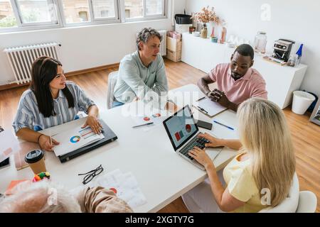 A multiethnic group of professionals works together at a coworking table. The image shows a Caucasian, Hispanic, and African American discussing over Stock Photo