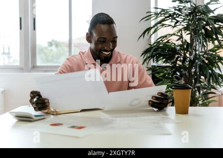 Cheerful African American man smiling while reviewing documents in a well-lit coworking space, surrounded by indoor plants and a coffee cup on his des Stock Photo