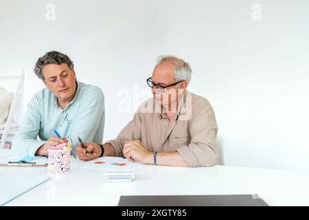 Two Caucasian men, of middle and older age, work together at a white table in a bright coworking space, engaging over work documents and using colorfu Stock Photo