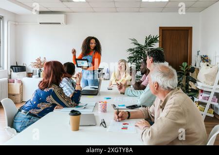 An African American woman stands, presenting on a laptop to a diverse group sitting at a table in a well-lit coworking space. Participants include Cau Stock Photo