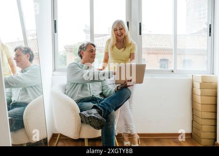 Senior Caucasian man sits on an armchair, holding a laptop with his partner, an Caucasian woman, standing beside him. Both are smiling and actively en Stock Photo