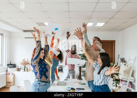 A joyful, multiethnic group of professionals celebrate by throwing papers in the air in a bright coworking office. Featuring male and female colleague Stock Photo