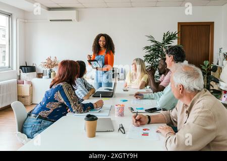 A multiethnic group of professionals engages in a collaborative session in a well-lit coworking space. An African American woman stands presenting wit Stock Photo