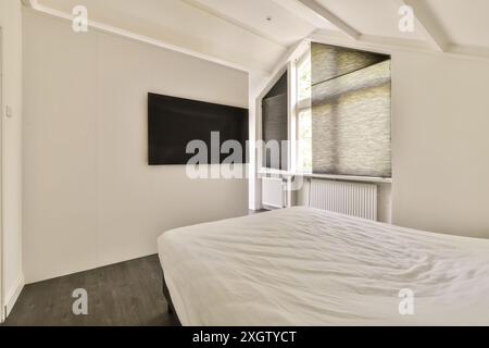 A modern bedroom features minimalist design with a large bed, wall-mounted TV, and natural light filtering through woven blinds in a Hilversumstraat a Stock Photo