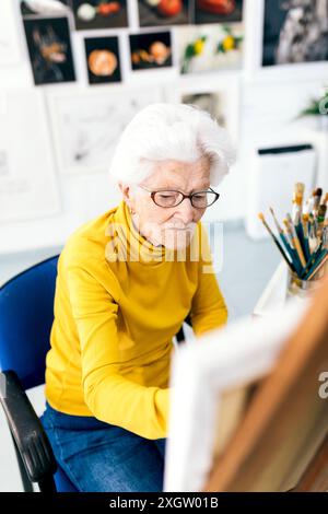 An elderly woman focuses intently on painting in a light-filled art studio, surrounded by brushes and art on the wall Stock Photo