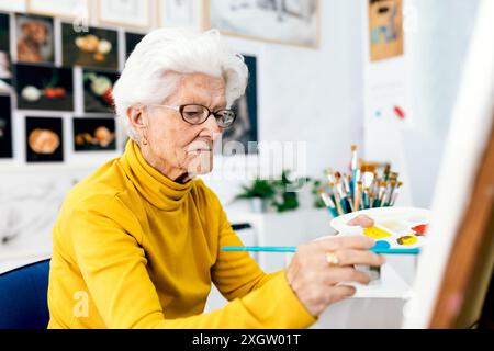 An elderly woman in a yellow sweater focuses intently on her canvas as she paints, surrounded by images and art supplies Stock Photo