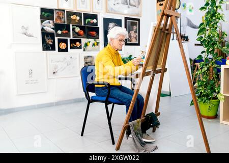 A senior woman in a yellow sweater focuses on painting on an easel in a vibrant art studio surrounded by various artworks Stock Photo