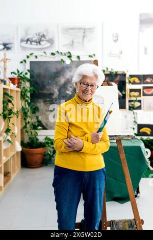 A cheerful elderly woman artist stands in her well-lit art studio, holding paint brushes, surrounded by lush plants and artwork Stock Photo