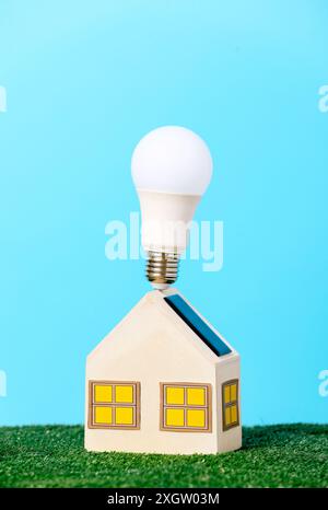 A mini wooden house model crowned with an eco-friendly LED bulb symbolizing sustainable housing solutions, placed on lush green grass with a vibrant b Stock Photo