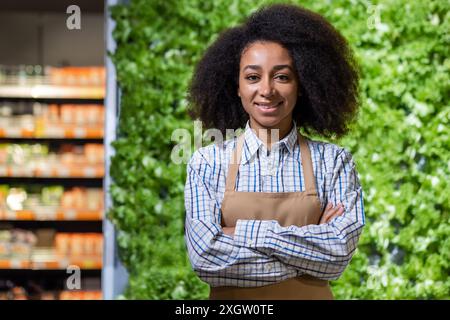 Confident young woman in apron standing in grocery store with fresh produce display in background. Ideal image for themes of retail, small business, entrepreneurship, and customer service. Stock Photo