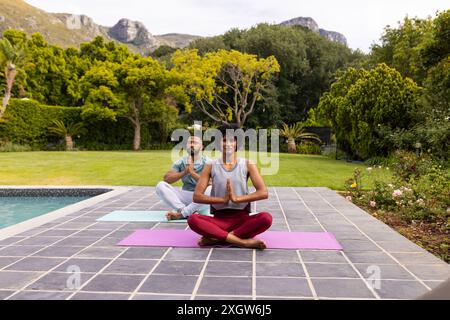 Biracial couple practicing yoga outdoors, with copy space. They enjoy a serene session by the pool surrounded by nature. Stock Photo
