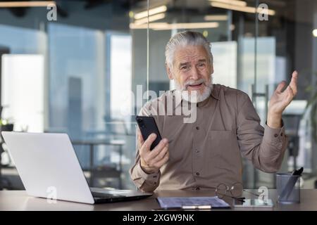 Elderly businessman expressing confusion while holding smartphone and looking at laptop. Scene set in modern office with glass walls. Concept of technology challenges, senior worker adaptation Stock Photo