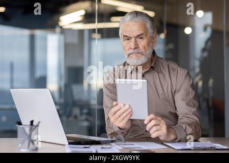 Senior businessman with beard using tablet seated at modern office desk with laptop, documents, and office supplies. Focused expression, professional attire, contemporary background Stock Photo