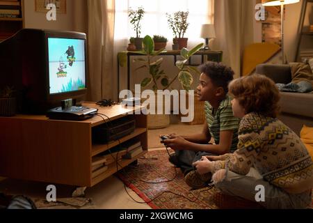 Two multiethnic boys engaged in playing a retro video game on an old television set, sitting on a carpeted floor with plants and home decor in the background Stock Photo