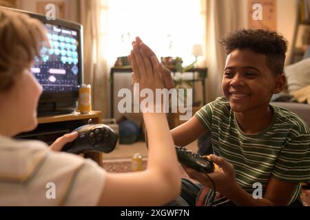 Over the shoulder shot of two diverse teen boys sitting in front of CRT TV with retro controllers in their hands Stock Photo