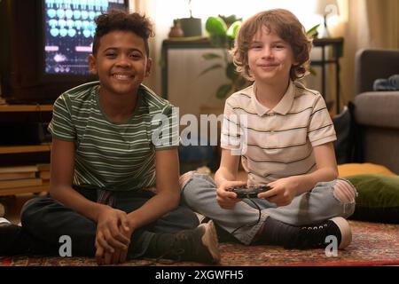 Portrait of two diverse boys smiling, looking at camera and sitting on carpeted floor while playing retro video games during free time at home Stock Photo