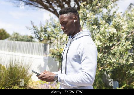 Focused african american man training outdoors, wearing earphones and using smartphone. Communication, jogging, fitness, exercise, wellbeing and healt Stock Photo