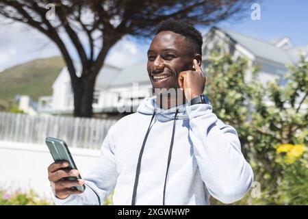 Happy african american man training outdoors, wearing earphones and using smartphone in sun. Communication, jogging, fitness, exercise, wellbeing and Stock Photo