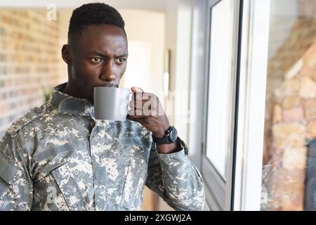 Thoughtful african american male soldier at home holding coffee and looking out of window. Depression, ptsd, military service, army, return, home, men Stock Photo