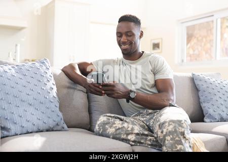 Happy african american male soldier sitting on couch at home using smartphone and smiling. Communication, military service, army, return, home, domest Stock Photo