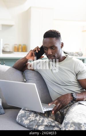 Focused african american male soldier at home using laptop and talking on smartphone, copy space. Communication, military service, army, return, home, Stock Photo
