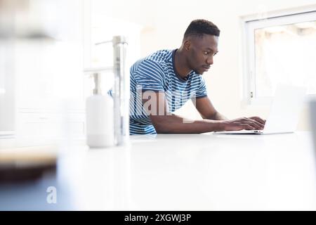 Thoughtful african american man using laptop leaning on worktop in sunny kitchen, copy space. Communication, refreshment, morning, domestic life and l Stock Photo
