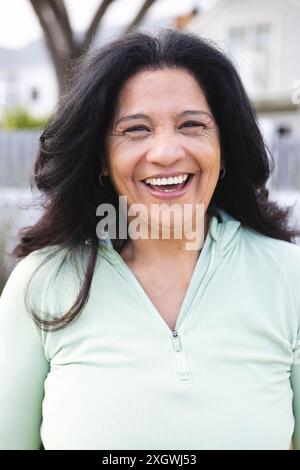 Vertical portrait of happy senior biracial woman with long black hair smiling outdoors. Wellbeing, health and active senior lifestyle, unaltered. Stock Photo