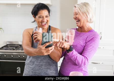 Happy diverse senior female friends drinking smoothies in kitchen, looking at smartphone laughing. Food, nutrition, friendship, domestic life and heal Stock Photo