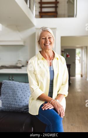 Portrait of happy senior caucasian woman sitting on arm of sofa smiling at home, copy space. Wellbeing, domestic life and senior lifestyle, unaltered. Stock Photo