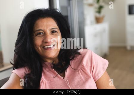 Portrait of happy senior biracial woman with long black hair sitting smiling at home. Wellbeing, domestic life and senior lifestyle, unaltered. Stock Photo