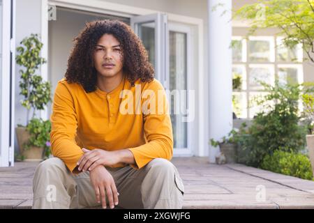 Young biracial man sits relaxed on a home porch. He exudes a casual confidence in a comfortable suburban setting. Stock Photo