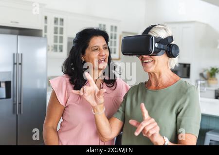 Two happy diverse senior female friends talking and using vr headset at home. Friendship, technology, digital interface, communication, retirement, do Stock Photo
