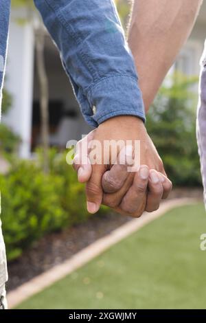 Diverse couple holding hands outdoors, with copy space. Their interlocked fingers symbolize love and unity. Stock Photo